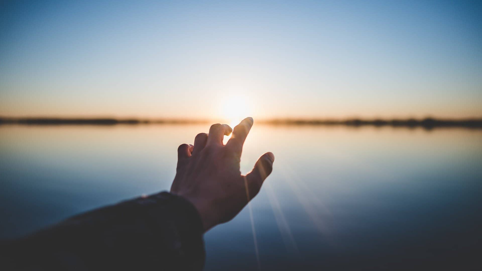 person extending their hand toward the sun near a body of water.