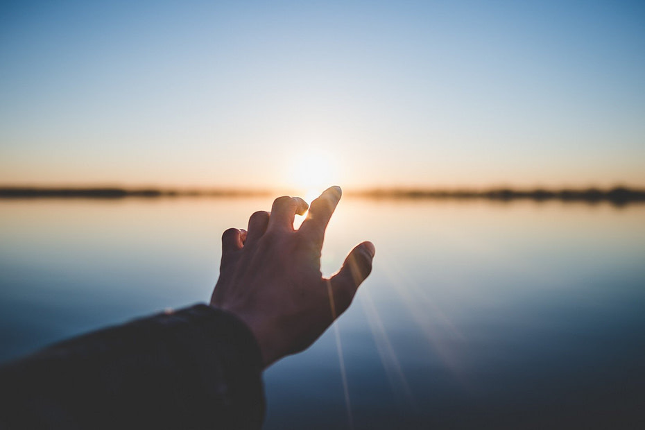person extending their hand toward the sun near a body of water.